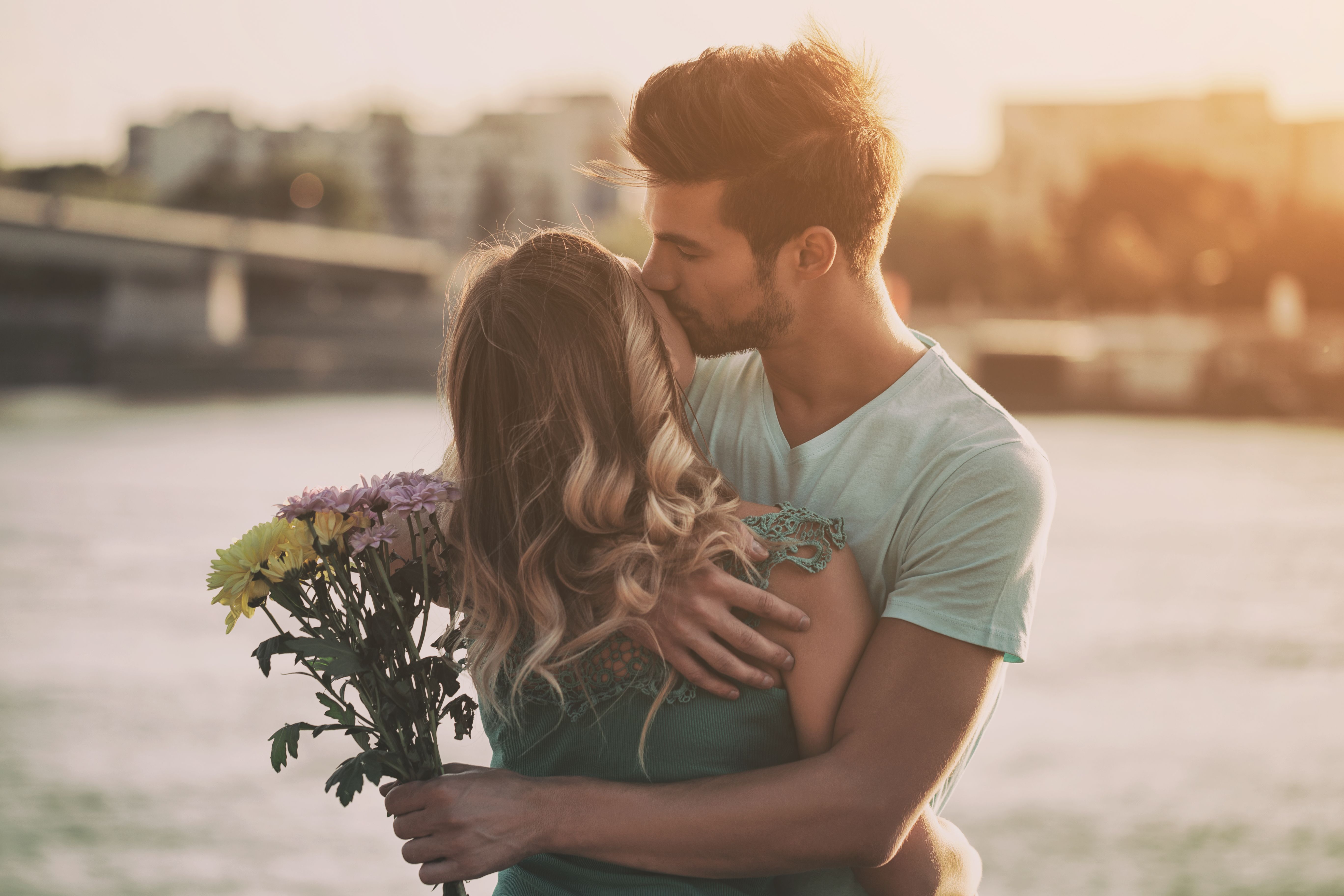 Jong koppel dat elkaar glimlachend omhelst in de stad, met een boeket bloemen in de hand.